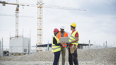 Baubesprechung einer Architektin mit zwei Bauarbeitern auf einer großen Baustelle (mit mehreren Baukränen im Hintergrund). Sie hält einen geöffneten Laptop in der Hand. Einer der Bauarbeiter schaut auf den Bildschirm.
