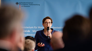 Bundesministerin Klara Geywitz begrüßt die Gäste mit einem Mikrofon in der Hand. Der Fotograf stand zwischen den Gästen und hat einige Hinterköpfe mitfotografiert.