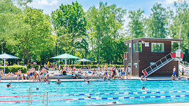 Schwimmbecken eines Freibades. Rechts am Bildrand der Aufsichtsturm der Rettungsschwimmer. Am Beckenrand und im Wasser viele Personen. Im Hintergrund grüne Bäume