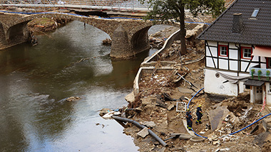 An einem Fluß ist die Steinbrücke stark beschädigt . Am rechten Bildrand steht ein ebenfalls stark bechädigtes Haus mit viel Schlamm und Müll auf dem Grundstück. Auf dem Grundstück sind 3 THW-Helfer aktiv.