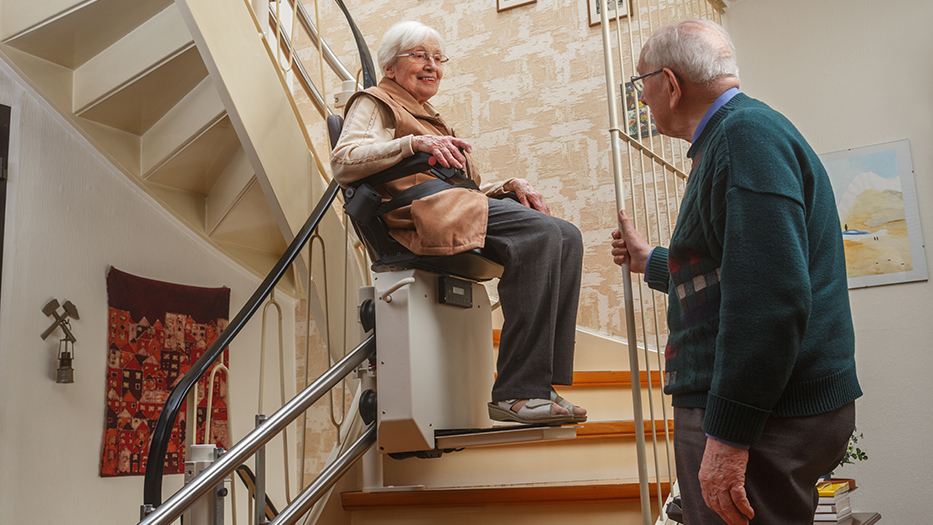 Eine ältere Frau in einem Treppenlift und einer älterer Mann, der ihr bei der Fahrt zusieht.