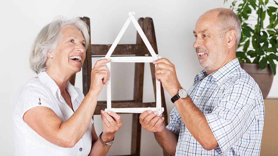 Älteres Ehepaar freut sich auf ihr neues Haus und haben mit einem Zollstock die Silhouette eines Hauses nachgestellt, welches sie zwischen sich halten und sich anlachen.