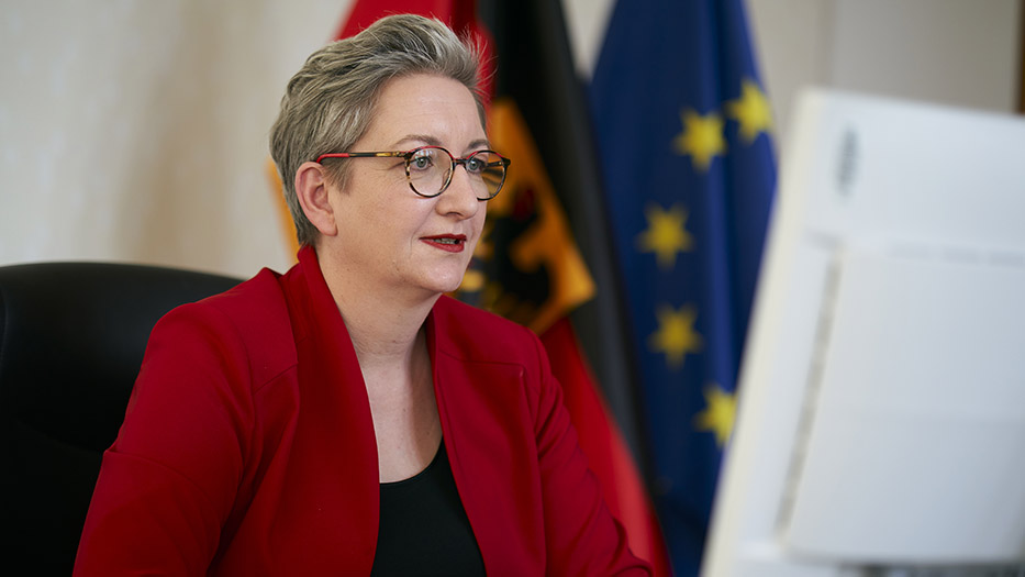 Bundesministerin für Wohnen, Stadtentwicklung und Bauwesen Klara Geywitz sitzt an Ihrem Schreibtisch vor einem Bildschirm und verfolgt eine Videokonferenz. Im Hintergrund die Bundesflagge und die Europaflagge.
