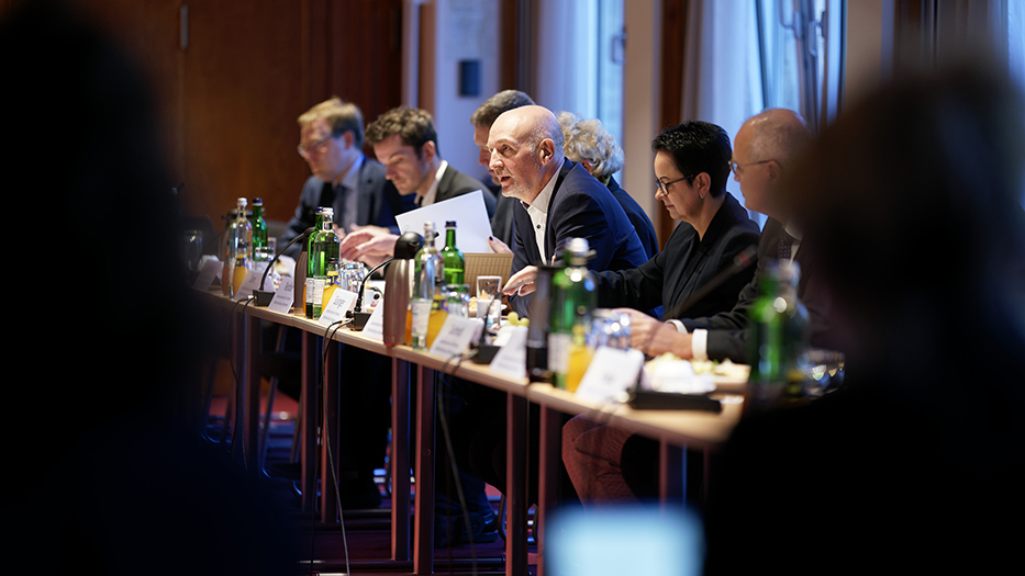 Durch zwei Teilnehmer hindurch fotografiert: Staatssekretär Bösinger sitzt neben weiteren Teilnehmerinnen und Teilnehmern am Konferenztisch
