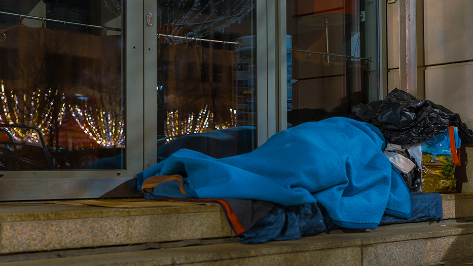 Vor dem Eingang eines Geschäfts liegt eine Person in Decken gehülllt. Im Glas der Eingangtür spiegeln sich Weihnachtslichter