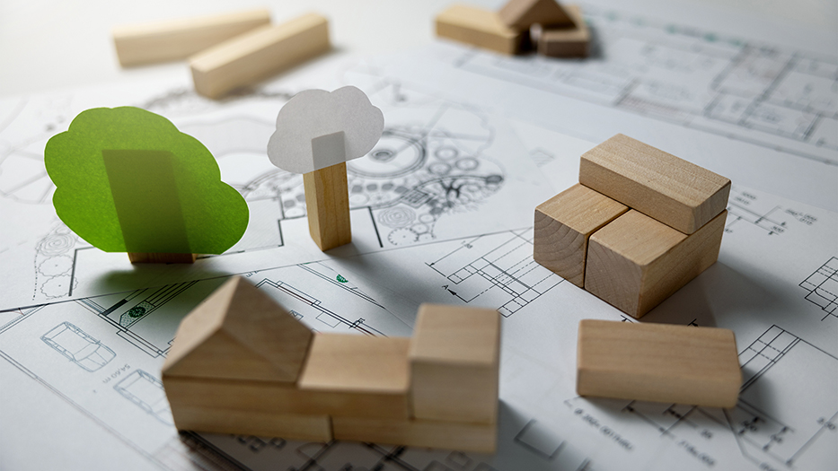 Auf einem Bauplan liegen Holzklötze und bilden ein Haus als Modell. Daneben sind Bäume aus Papier. Es wird den enerneuerbaren Materialien der umweltfreundliche Bau symbolisiert.
