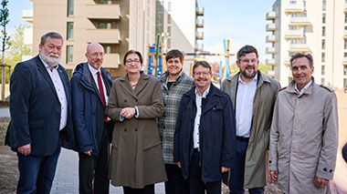 Gruppenfoto mit insgesamt 7 Personen, Klara Geywitz steht mittig. Im Hintergrund sind neu gebaute Mehrfamilienhäuser und ein Spielplatz zu sehen.
