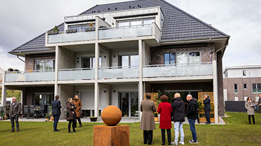 Die Ministerin betrachtet mit mehreren Personen ein modernes Mehrfamilienhaus mit Balkonen und einer Grünanlage.
