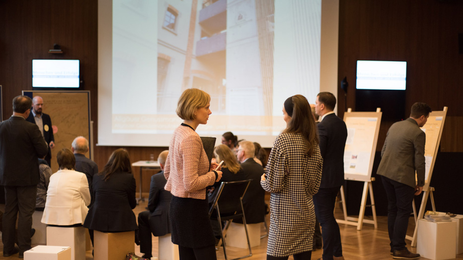 Archivbild: AUfnahme von der Veranstaltung 2019. Teilnehmerinnen und Teilnehmer unterhalten sich. Auf einer Leinwand im Hintergrund ist ein Wohnkomplex abgebildet.
