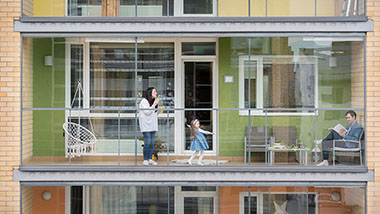 Aufnahme von einem Balkon eines Mehrfamilienhauses, der "Wintergarten" angelegt ist. Auf dem Balkon sind eine Mann im Stuhl. sowie eine stehende Frau mit Kind.