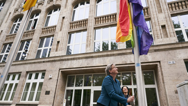 Bundesministerin Klara Geywitz am Fahnenmast vor dem Bauministerium. Sie hisst die Regenbogenflagge, die schon hoch über ihr hängt. BM'in Geywitz und die Parlamentarische Staatssekretärin Cansel Kiziltepe schauen hoch zur Flagge.