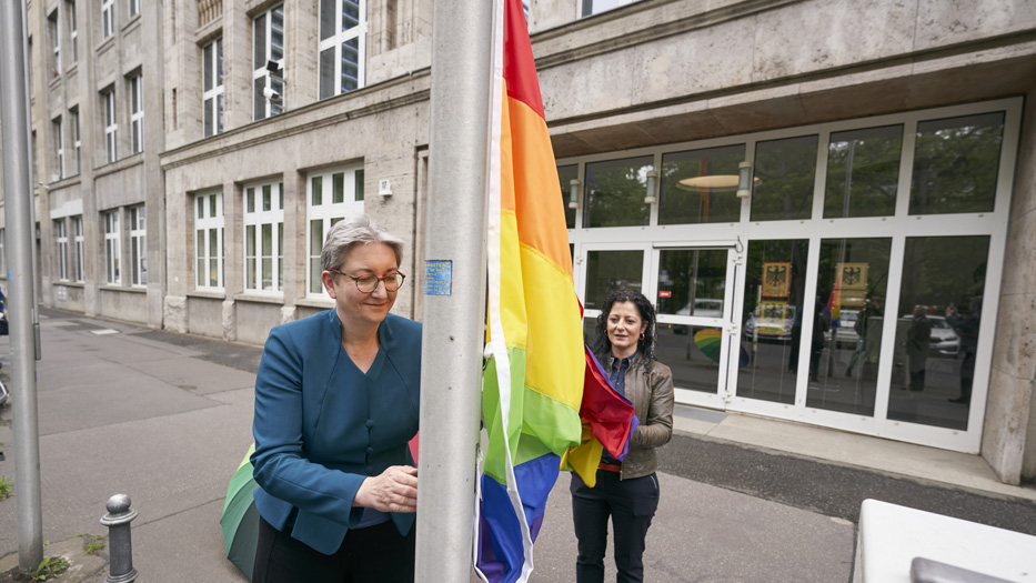 Bundesministerin Klara Geywitz am Fahnenmast vor dem Bauministerium. Sie hisst die Regenbogenflagge. Die Parlamentarische Staatssekretärin Cansel Kiziltepe ist ihr dabei behilflich, sodass die Flagge nicht auf dem Boden liegt.