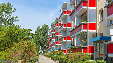 Symbolbild für genossenschaftliches Wohnen. Mehrfamilienkomplex mit einer Grünanlage. Die Balkone sind in rot abgesetzt.