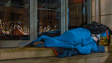 Vor einem geschützten Eingang zu einem Bürogebäude liegt eine Person in einem Schlafsack.